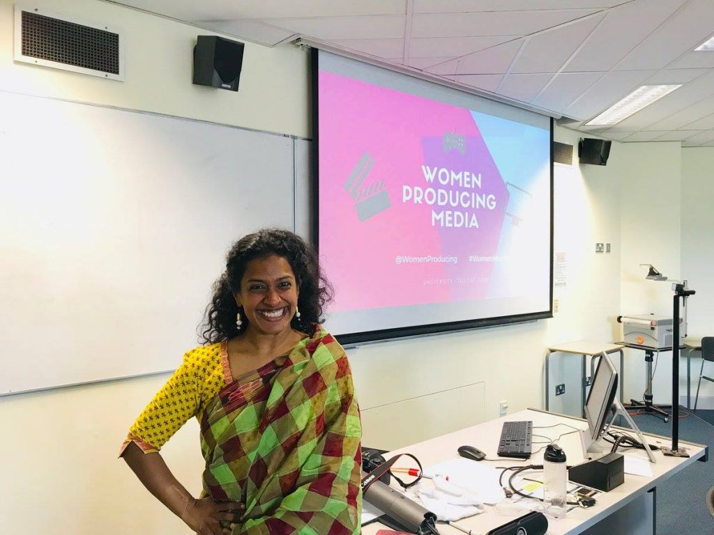 Joanna Dukkipati from the waist up, stands iwth one hand on her hip, in front of a desk and a large screen with the name of the event projected onto it.